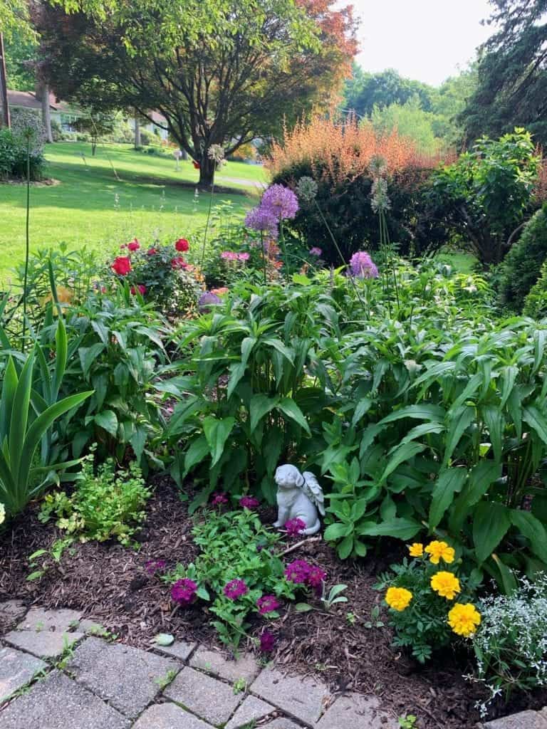 A lush garden with colorful flowers, green shrubs, and a small white angel statue near the center. A stone path borders the garden, and large trees and lawn are visible in the background.