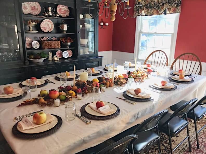 A dining table set for a meal with white plates, apples, candles, and autumn-themed decorations features a beautiful DIY table topper. The room is warmly lit with natural light and has a dark china cabinet with dishes in the background.