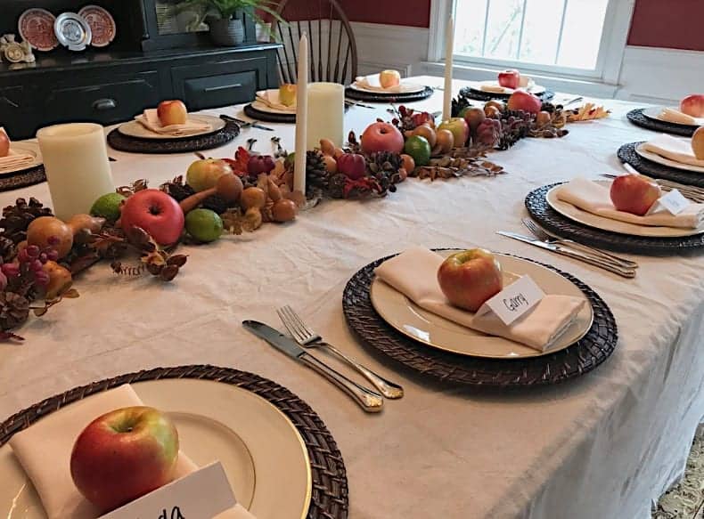 A dining table set for a meal with white plates, woven chargers, napkins, apples, name cards, and silverware. A diy table topper made of fruit, leaves, and candles runs down the middle as a stunning centerpiece.