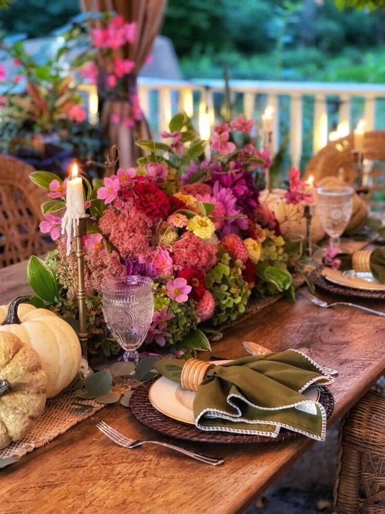 A rustic outdoor dining table is adorned with an elaborate, colorful floral centerpiece featuring pink, purple, and red blooms. Embracing a fall harvest table theme, it's set with woven placemats, elegant green napkins, and crystal wine glasses. A lit candle and decorative pumpkin accentuate the setup.