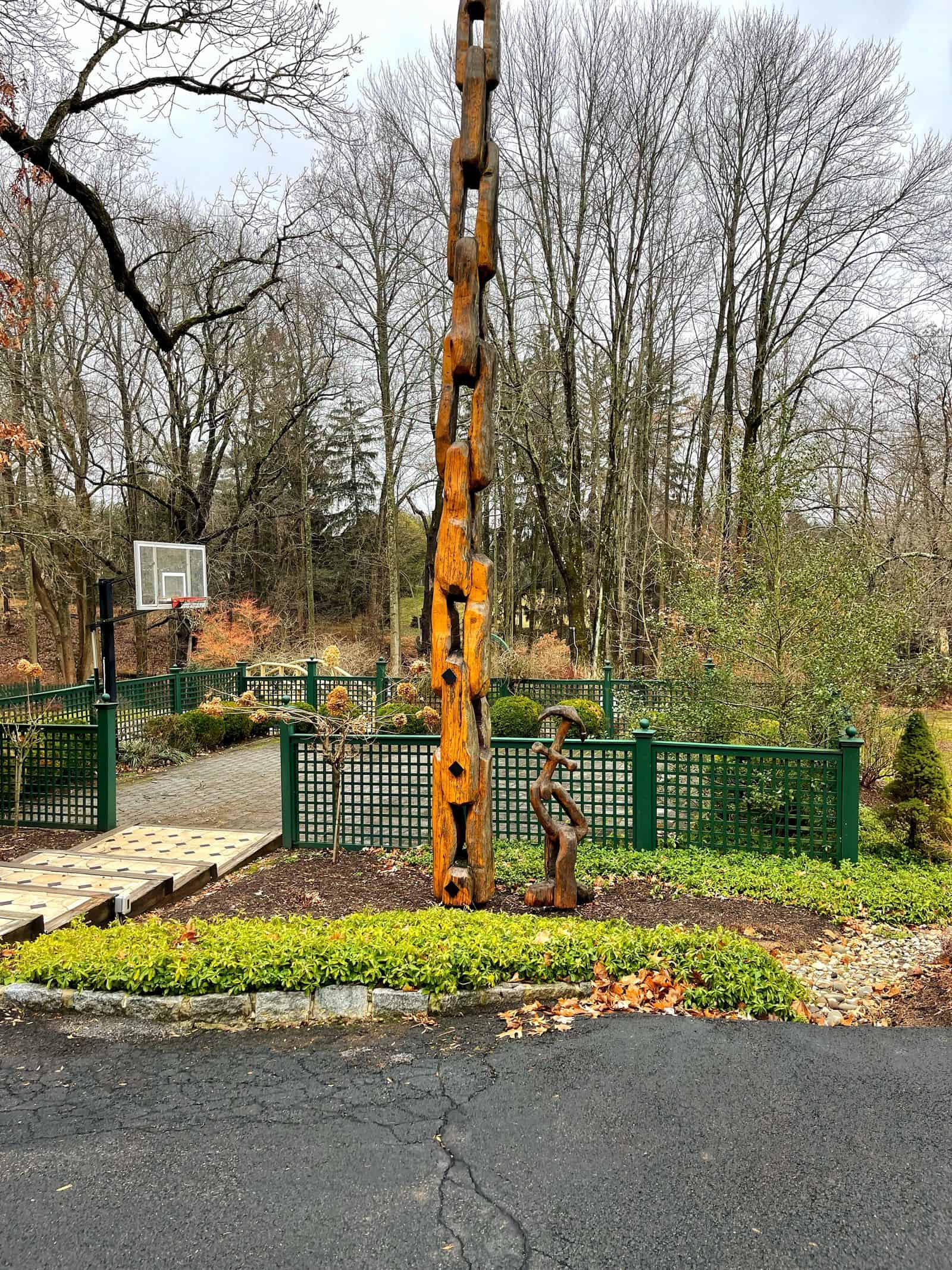 A tall, carved wooden sculpture stands in a landscaped potager garden before green fencing and bare trees; a basketball hoop is visible in the background on a cloudy day.