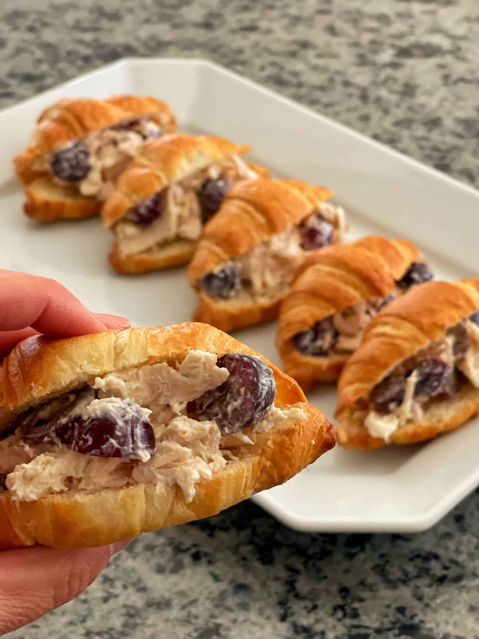A hand holds a small croissant sandwich filled with chicken salad with grapes. More similar mini croissant sandwiches, each bursting with chicken salad with grapes, are arranged on a white tray in the background on a speckled countertop.