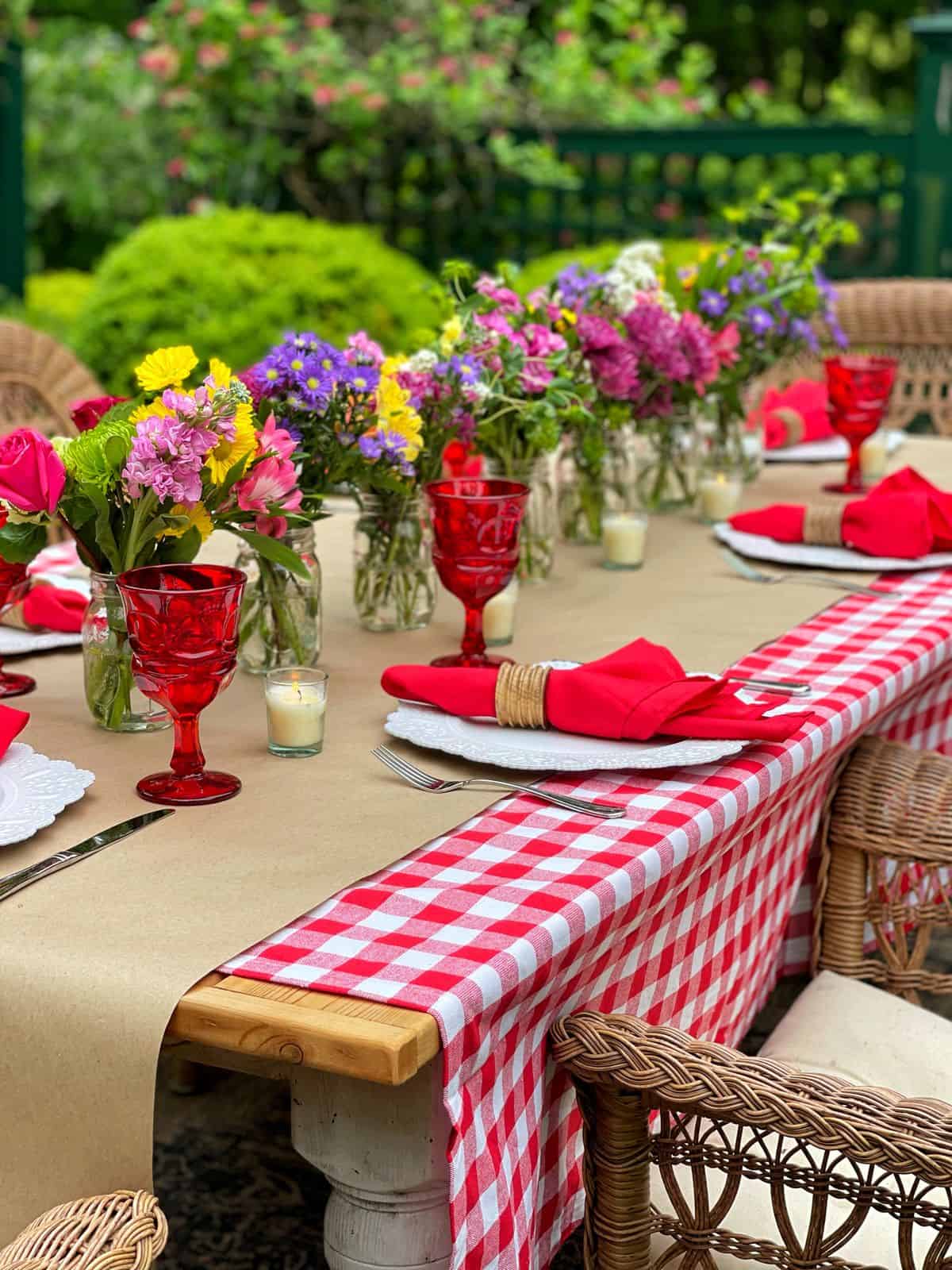 A picnic table set outdoors with a red and white checkered tablecloth, red glassware, red napkins, and jars of colorful flowers is ready for a festive low country boil, surrounded by wicker chairs and a lush green, leafy background.