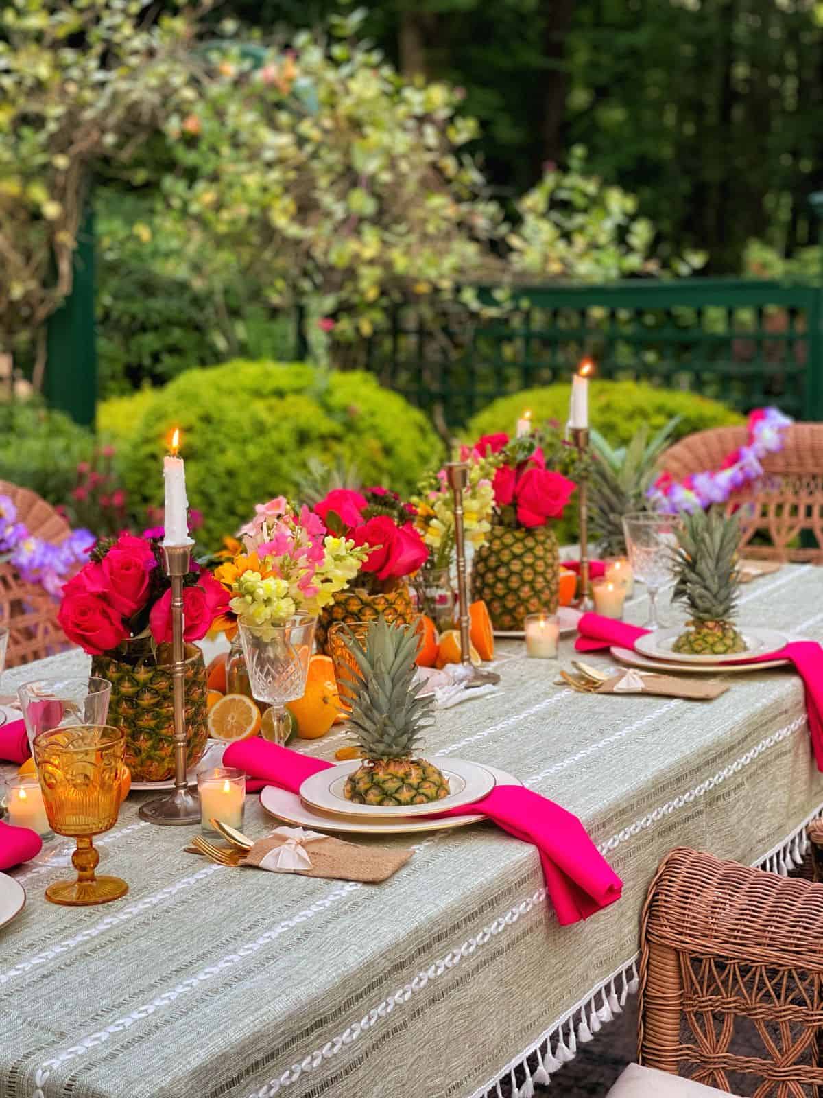 A festive outdoor table is set for a Hawaiian theme party with pineapples, bright pink flowers, candles, citrus fruits, gold cutlery, and pink napkins. Wicker chairs and lush green plants surround the elegant, colorful tablescape.