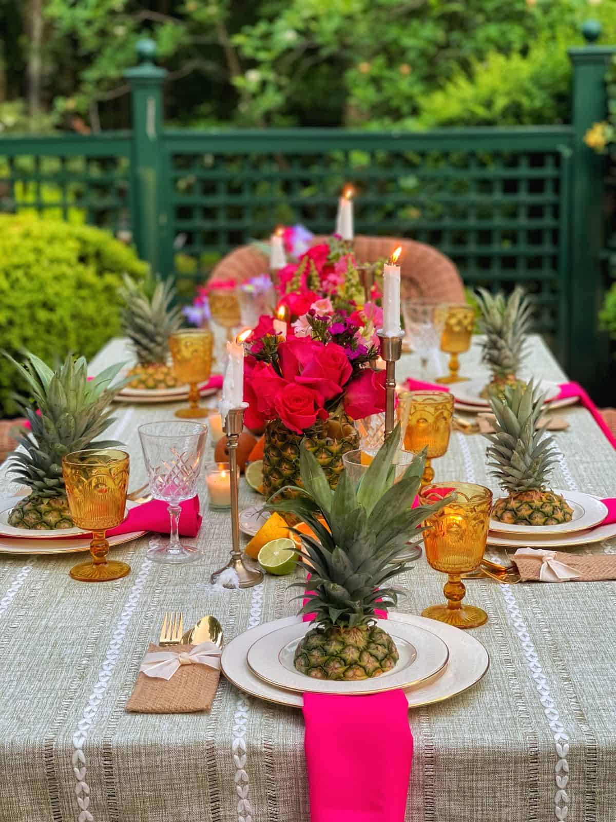 A colorful outdoor table set for a meal, featuring pineapples as centerpieces, pink flowers, amber goblets, candles, white plates, and bright pink napkins on a green tablecloth—perfect for those seeking Hawaiian party ideas in a garden setting.