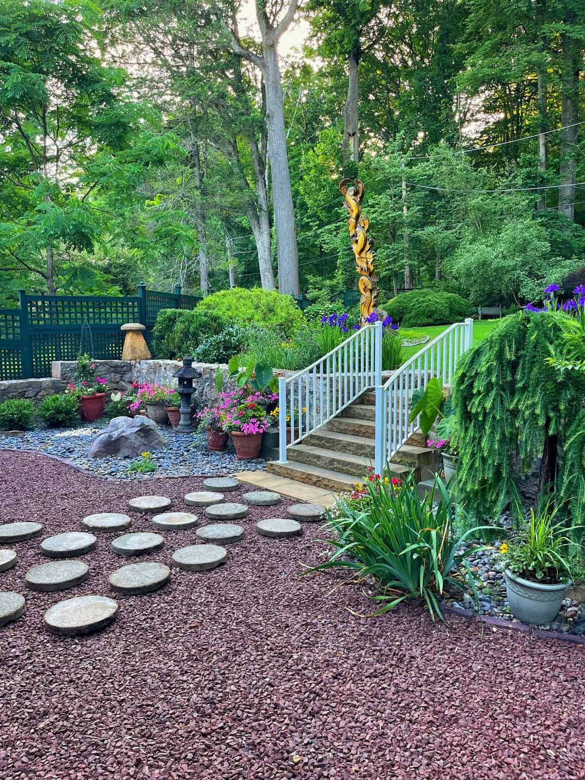A landscaped backyard zen garden with a stone path, white steps leading to a small bridge, lush greenery, potted flowers, a tall carved wooden sculpture, and a forested background.