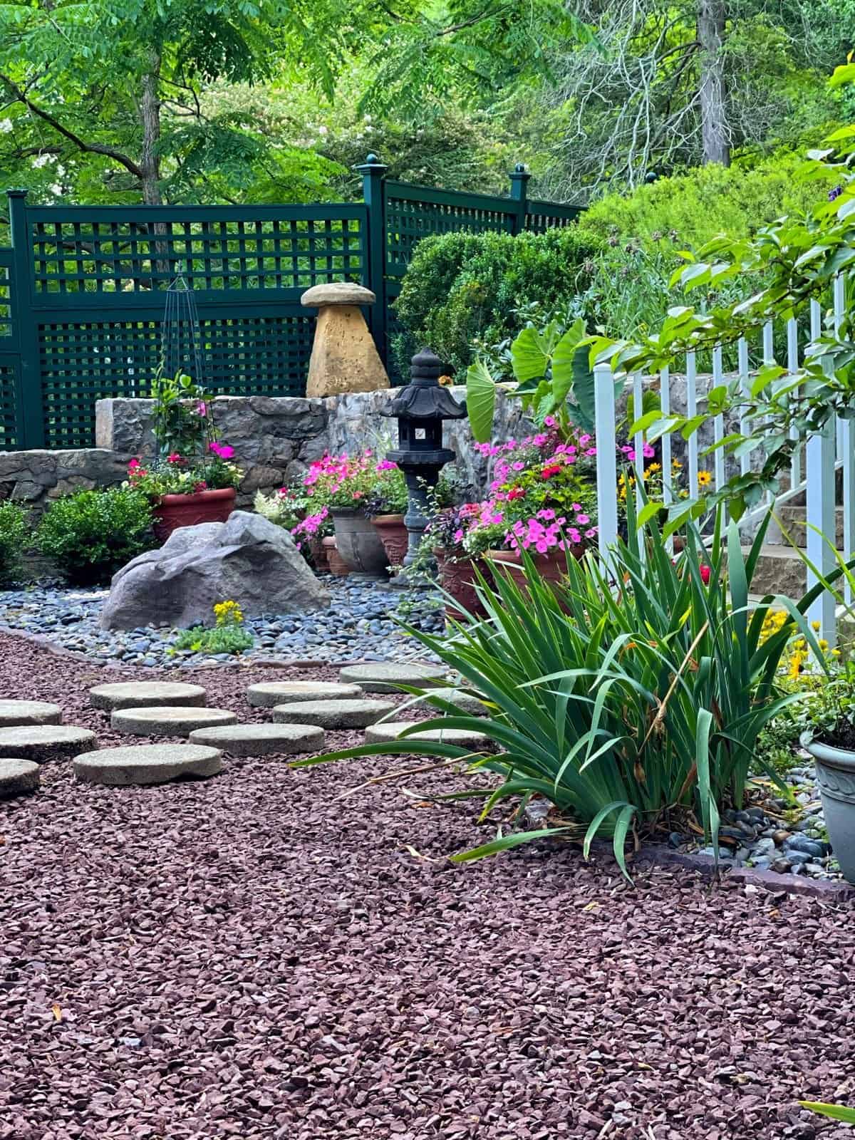 view of the zen garden with round stepping stones, container gardens filled with supertunia bubble gum pink petunias bubblegum pink, garden statues, lots of hardscaping and a green garden fence with a stone wall and white railings that lead to the backyard.