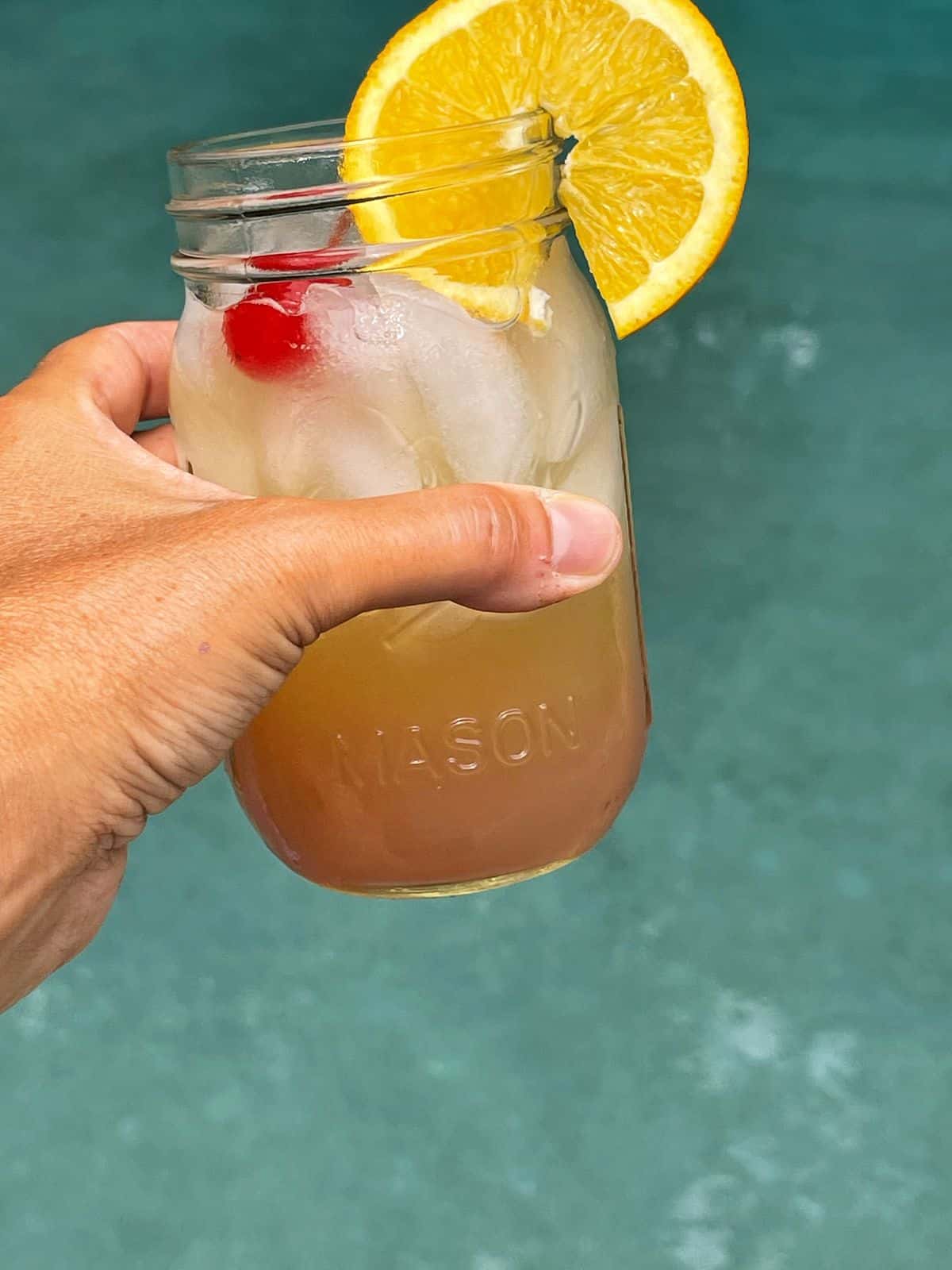 A hand holding a mason jar filled with an iced malibu sunset cocktail, garnished with a slice of orange and a cherry, against a blurred blue background that looks like a swimming pool.