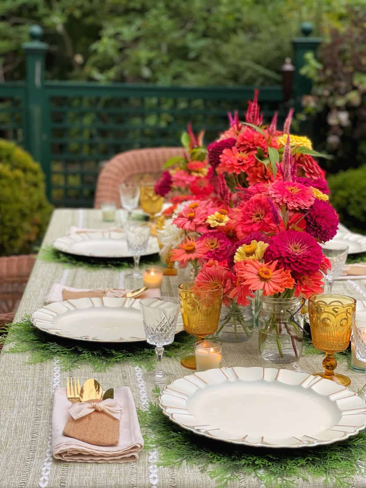 A beautifully set outdoor dining table perfect for a garden party features white scalloped plates, gold flatware wrapped in napkins, amber glasses, votive candles, and vibrant pink and orange flower arrangements amid lush greenery.