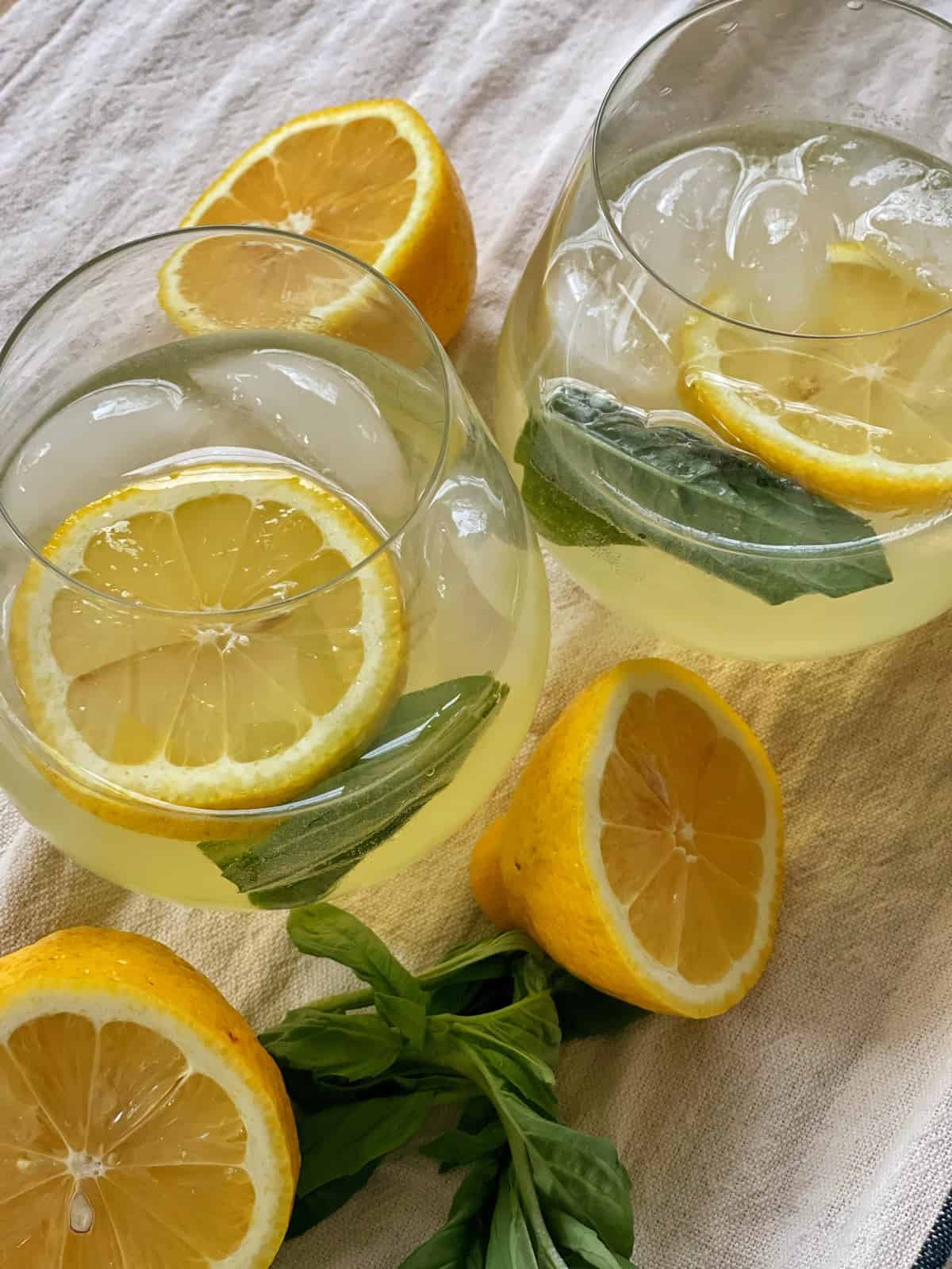 Two glasses of iced lemon basil drink, inspired by a hugo spritz, with lemon slices and basil leaves, placed on a light cloth, surrounded by halved lemons and fresh basil.