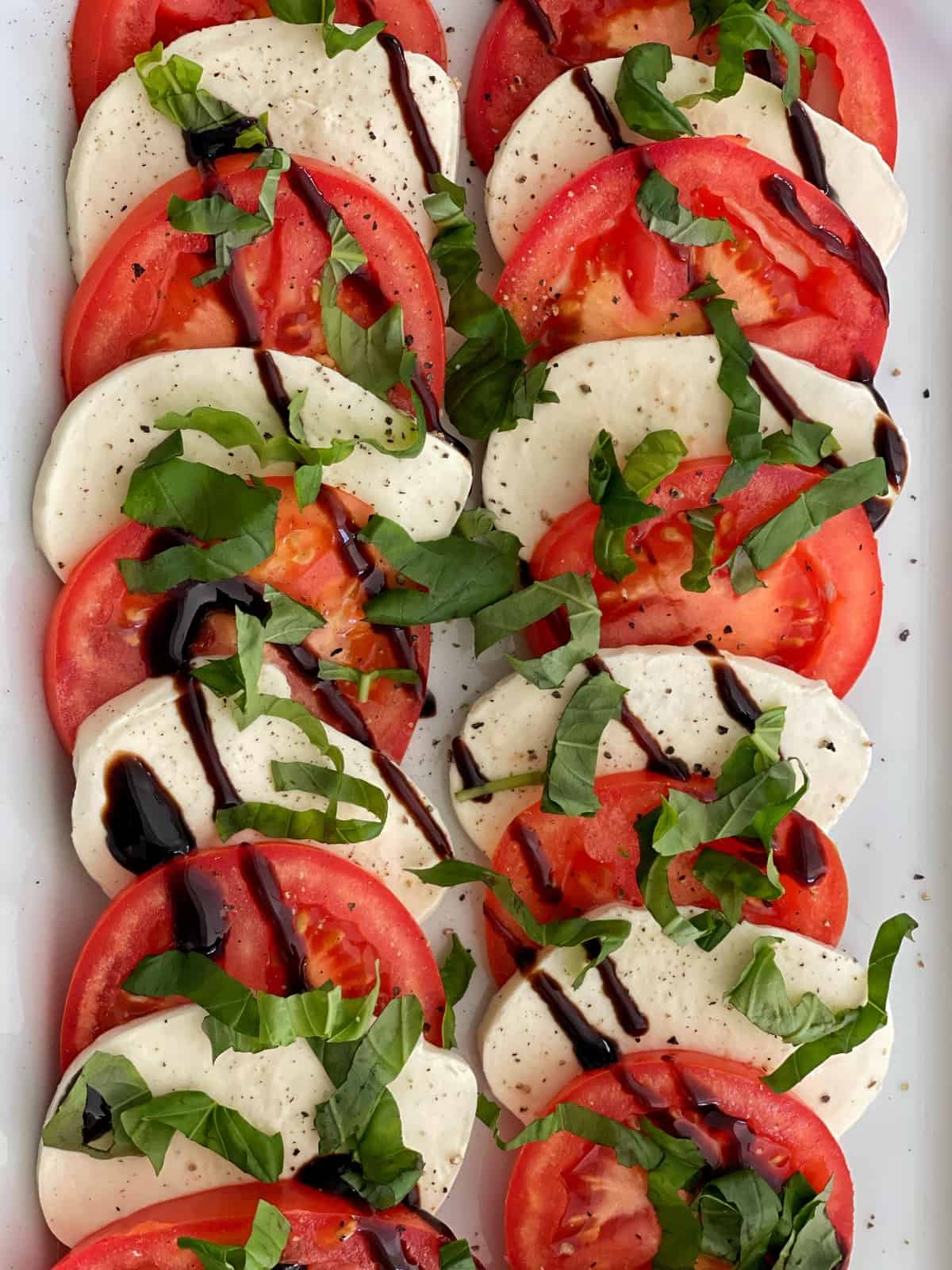 Caprese salad features sliced fresh tomatoes, mozzarella cheese, and basil leaves arranged in alternating layers on a platter, then drizzled with balsamic glaze and sprinkled with black pepper.