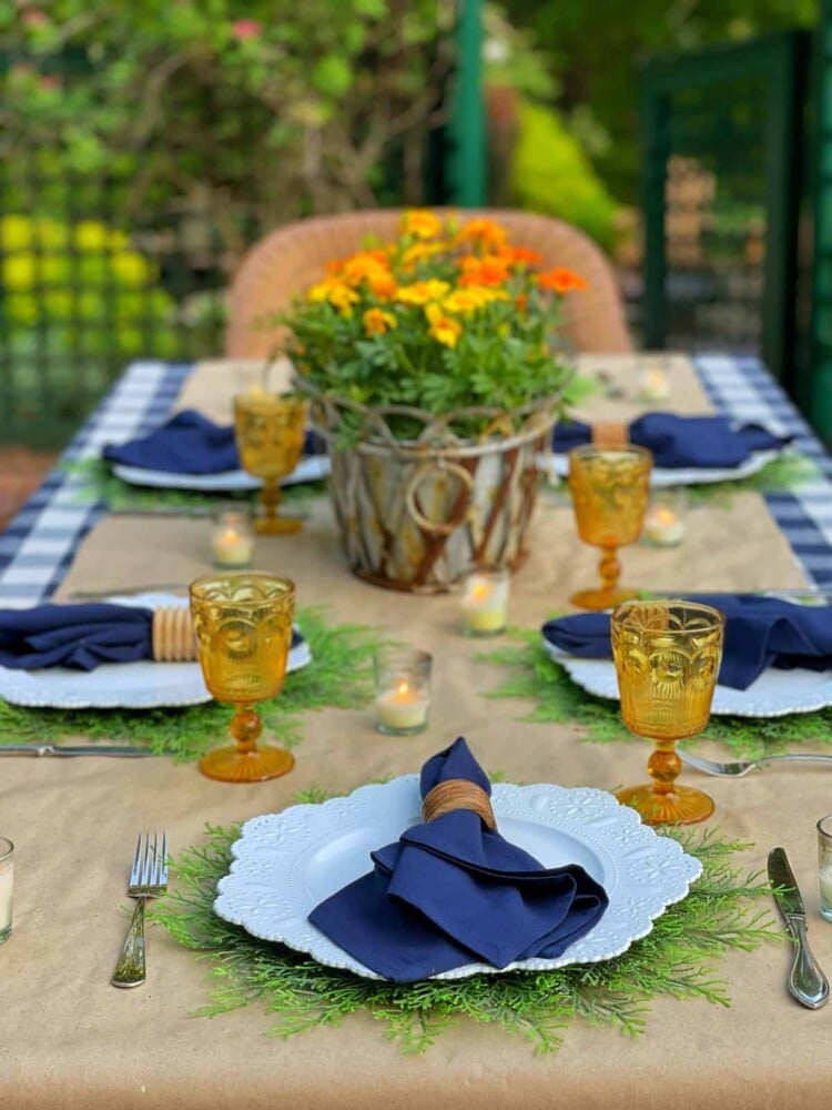 An inviting summer bbq table set with white plates, navy blue napkins, gold glasses, and small candles. A rustic basket filled with orange flowers serves as the centerpiece while greenery decorates the table runner.