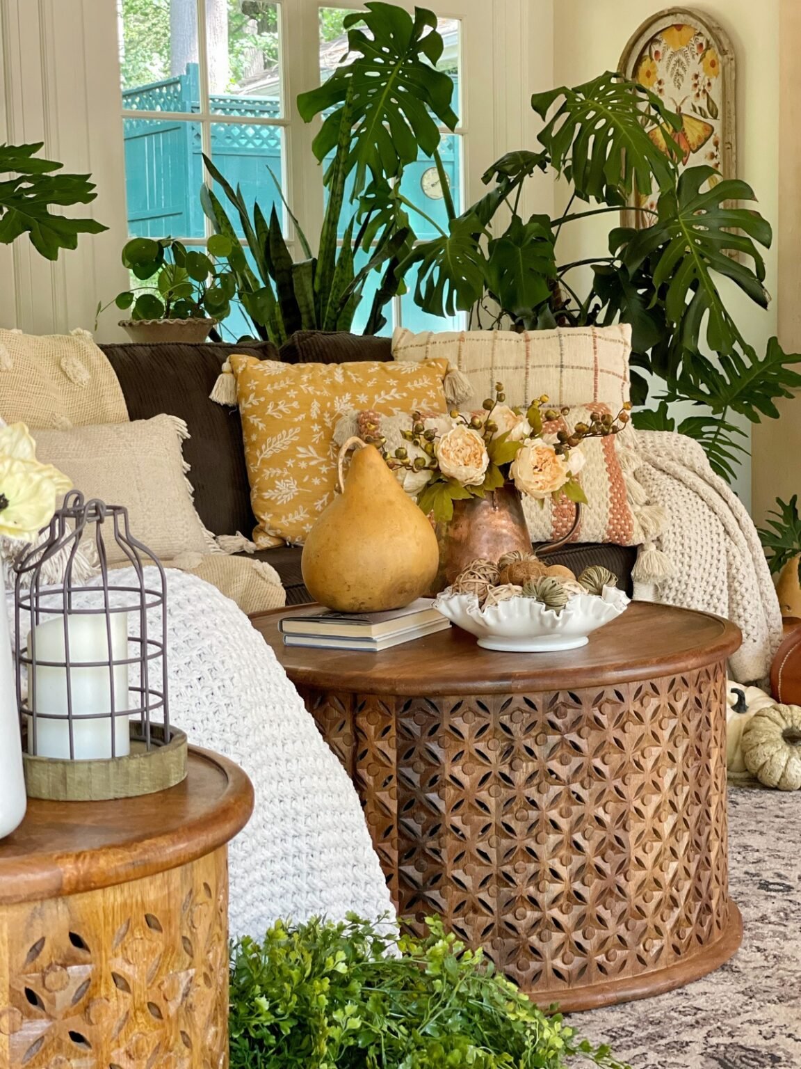 A cozy family room scene with a dark sofa adorned with white and mustard-colored pillows and a chunky knit throw, perfect for early fall decor ideas. A carved wooden coffee table holds a vase with flowers, a gourd, a book, and decorative items. Large green plants are in the background.