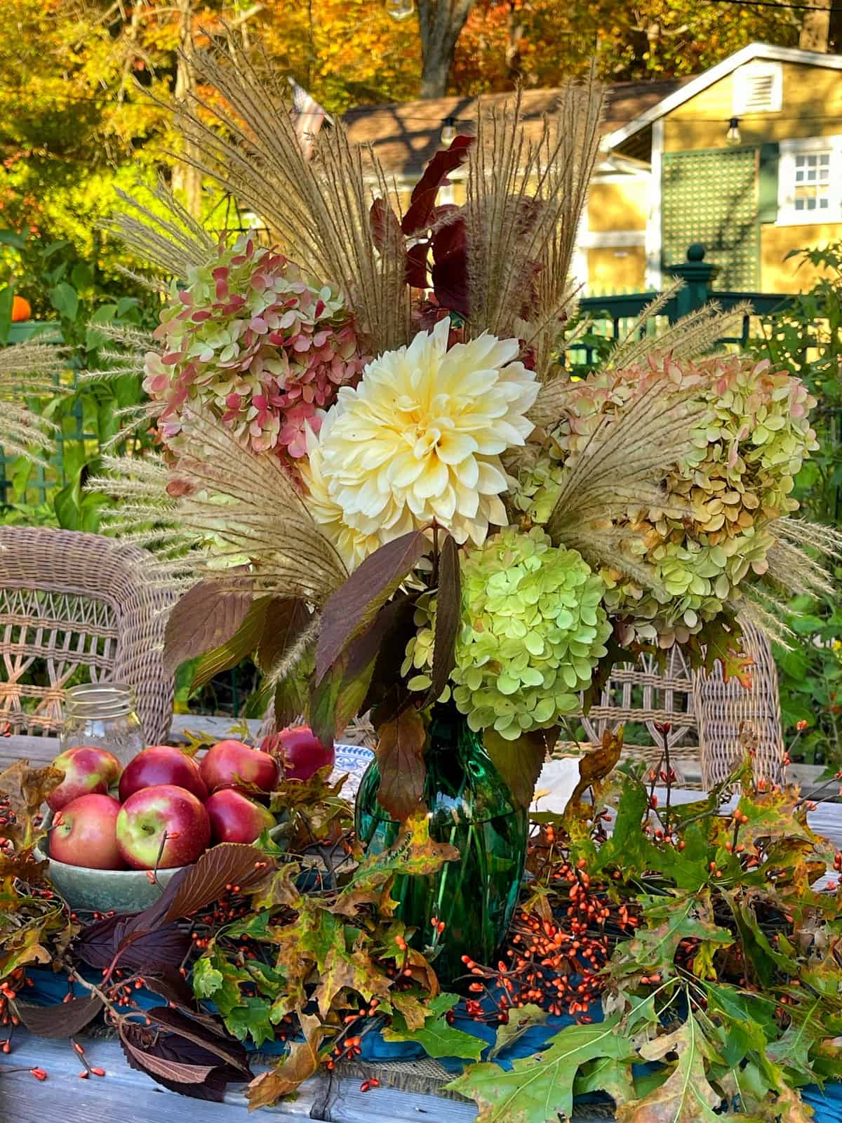 A vibrant autumn centerpiece sits on a wooden table, featuring a green vase filled with a mix of dahlias, hydrangeas, and dried grasses. A bowl of red apples and scattered fall leaves and berries add to the festive seasonal decor. A wicker chair and a cottage are in the background.