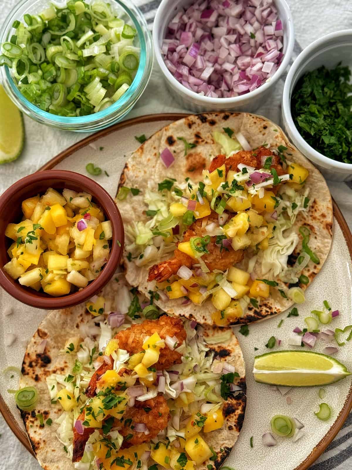 Three coconut shrimp tacos topped with mango salsa, cabbage, and herbs on a plate, accompanied by bowls of chopped onions, green onions, and limes on a textured white tablecloth.