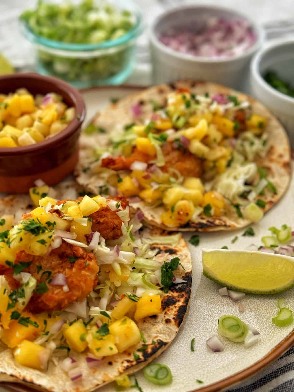 Two coconut shrimp tacos garnished with mango salsa, shredded cabbage, diced onions, and chopped cilantro on a white plate, served with lime wedges and additional toppings in the background.