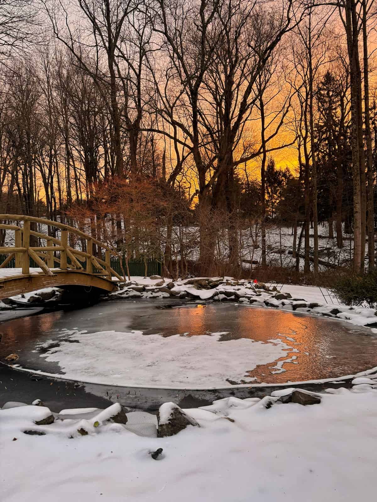 A snow-covered pond with patches of ice sits beside a wooden bridge, surrounded by leafless trees. The sky glows orange at sunset, casting a warm reflection on the icy water.