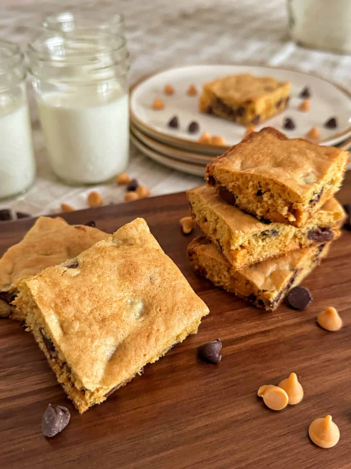 Several square blondie brownies with chocolate and butterscotch chips are stacked on a wooden board. Extra chocolate and butterscotch chips are scattered around, with glasses of milk and plates in the background.