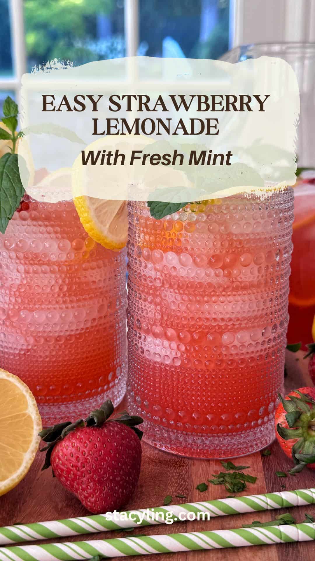 Two textured glasses of pink strawberry lemonade with lemon slices and fresh mint, surrounded by strawberries, lemon halves, and green-striped straws on a wooden table. Text on image reads, "Easy Strawberry Lemonade Recipe With Fresh Mint.