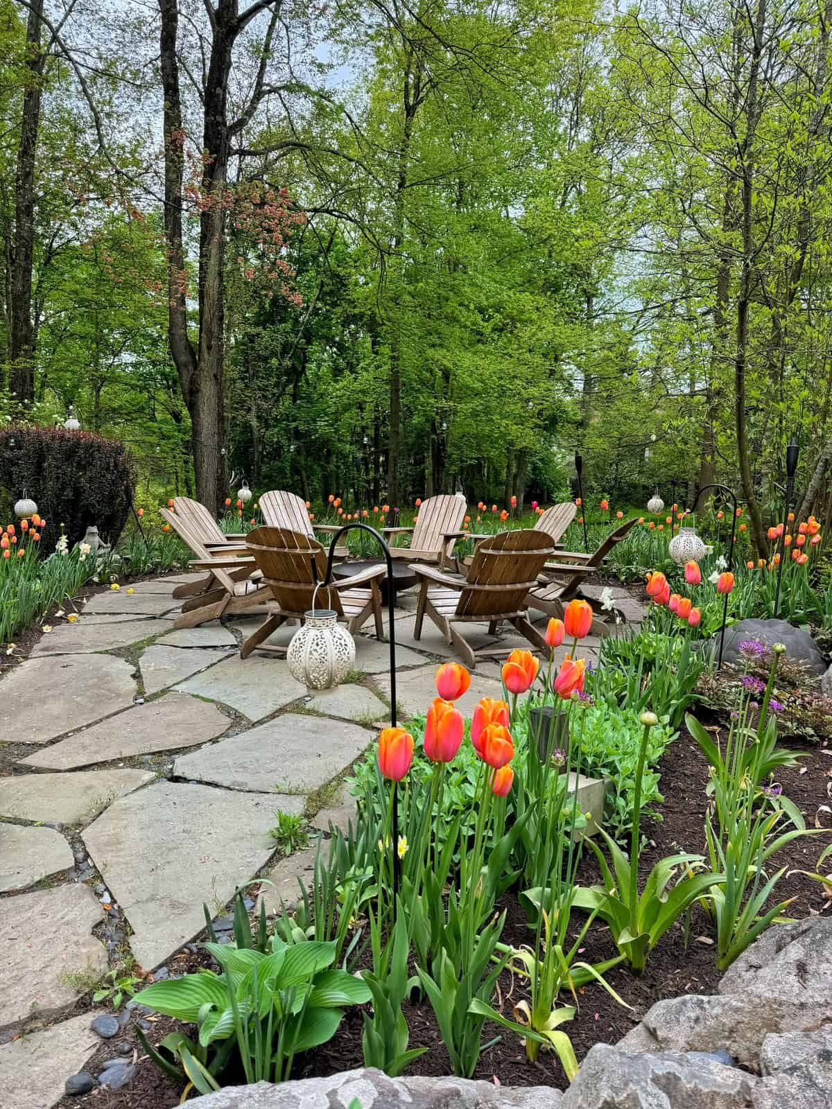 A serene garden featuring a stone pathway leading to a circle of wooden adirondack chairs surrounded by vibrant tulips and lush greenery.
