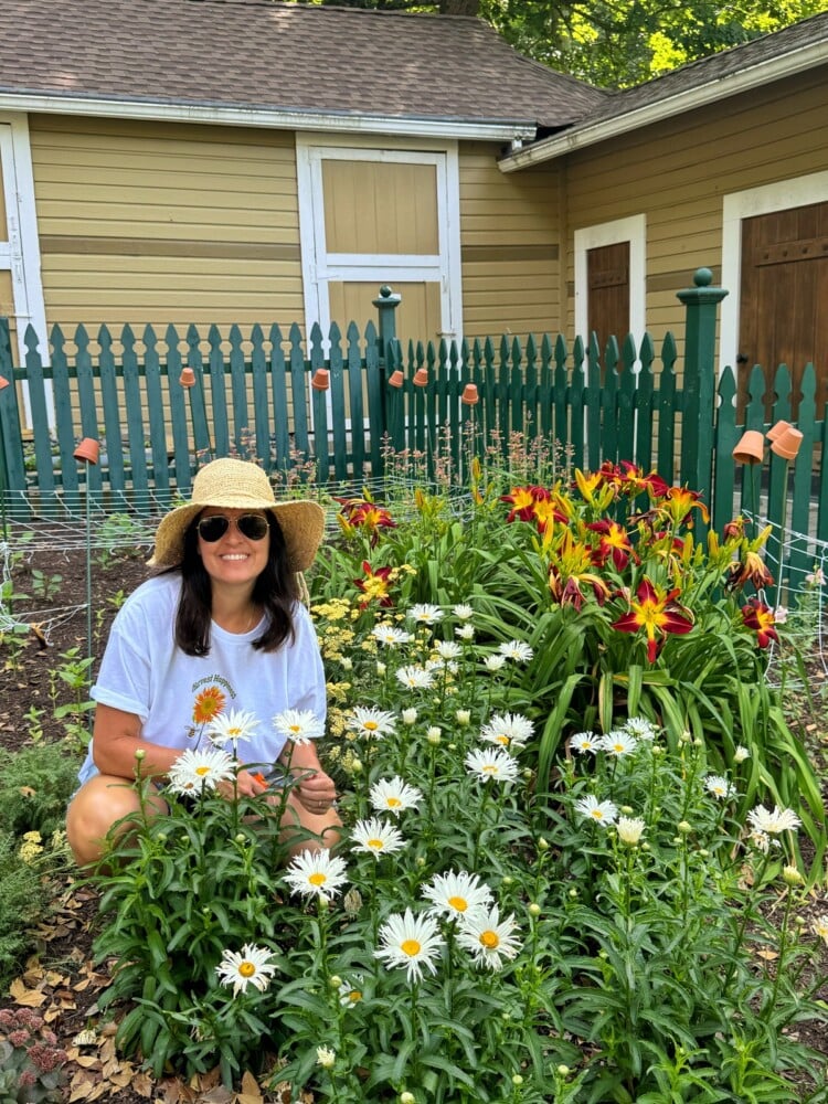 A woman (stacy ling) wearing a straw hat and sunglasses kneels in a garden filled with blooming flowers, including white shasta daisies and vibrant red and yellow lilies. She is smiling, dressed in a white shirt, while deadheading flowers to ensure their vibrancy. A picket fence and a yellow house stand in the background.