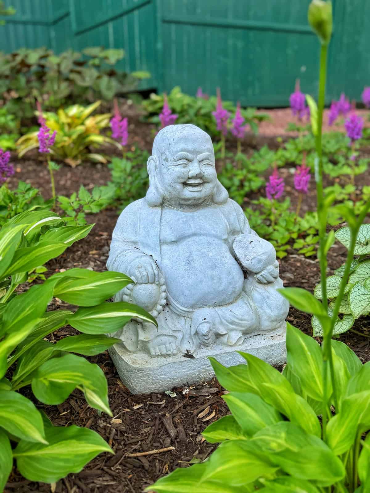 A smiling stone Buddha statue is sitting in a peaceful garden surrounded by green plants and vibrant purple flowers. The backdrop features a green wooden fence. The atmosphere is serene and tranquil.