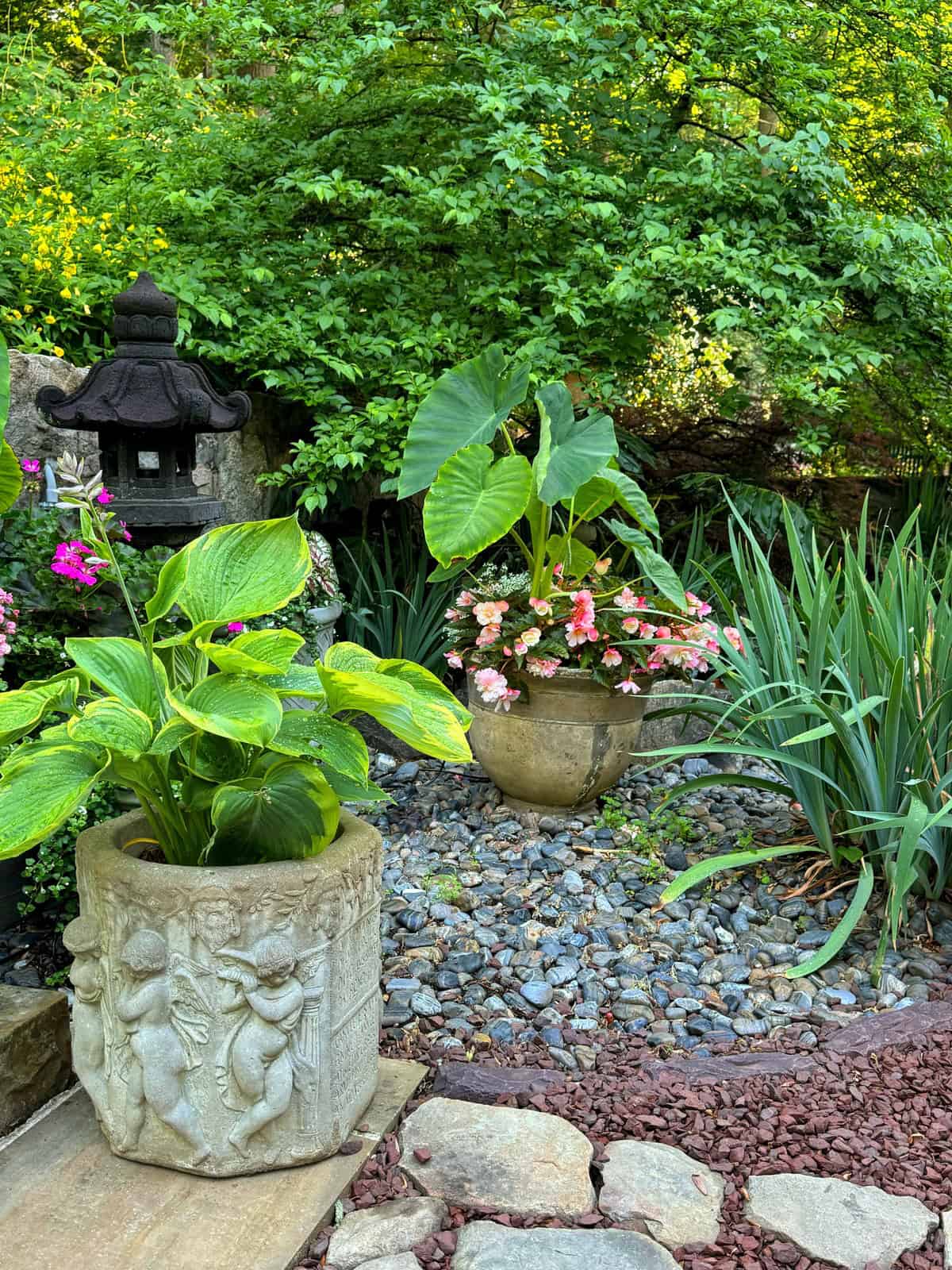 A serene garden scene featuring ornate stone planters filled with lush green plants and colorful flowers. Behind the planters, there's a Japanese-style lantern set amidst greenery. The ground is covered with small rocks and mulch, enhancing the tranquil ambiance.