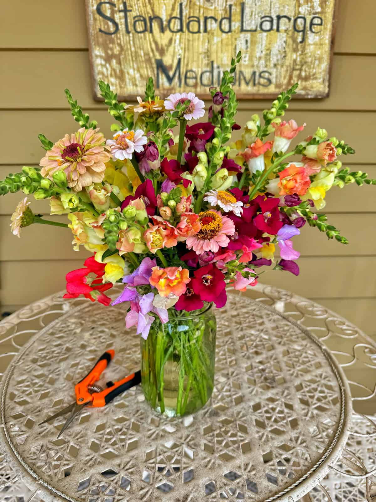 A mixed arrangement of colorful flowers in a clear glass jar rests on an ornate metal table. Small orange garden shears are placed to the left of the jar. A weathered sign with partially readable text hangs in the background.
