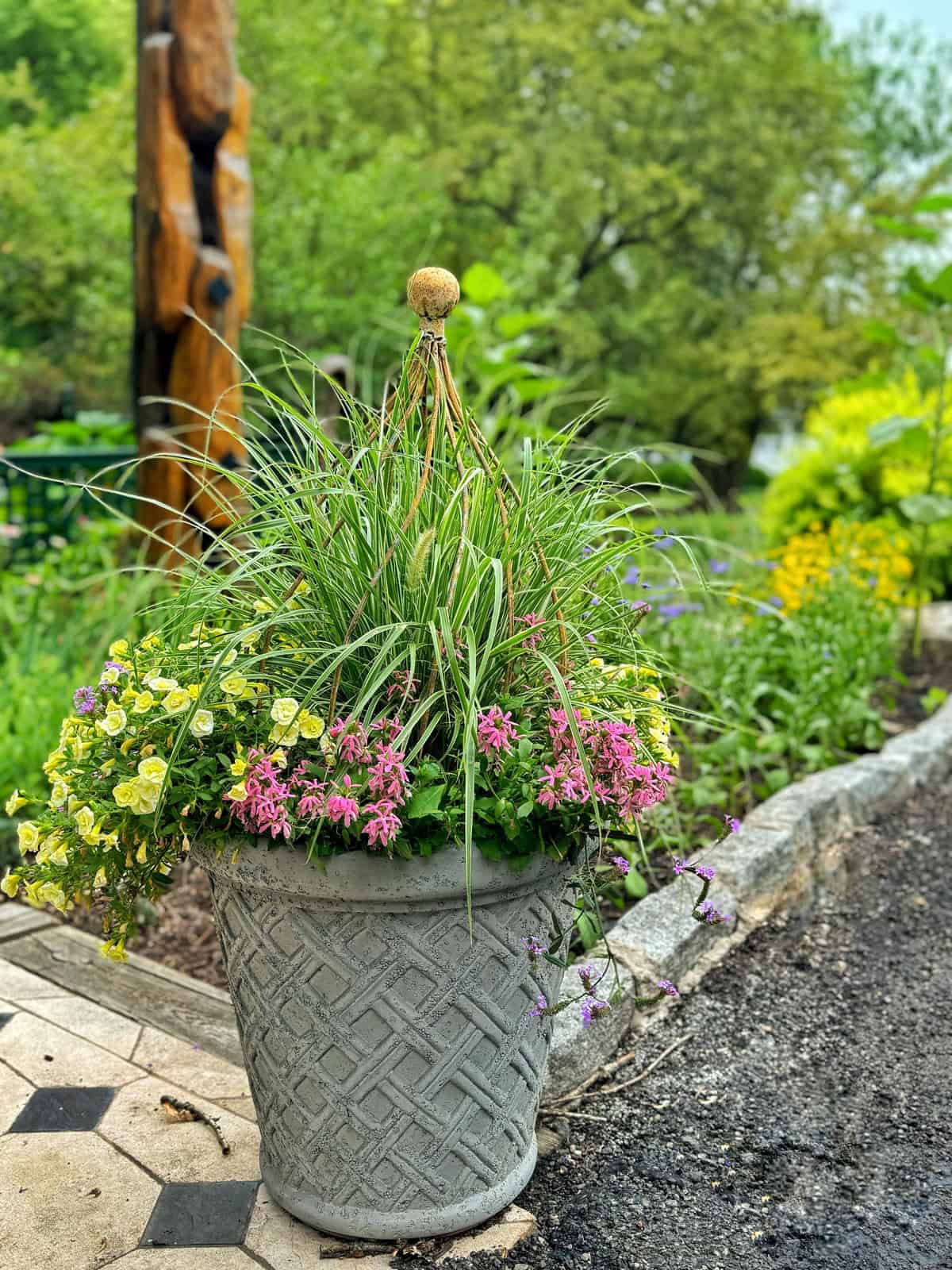 A large, decorative stone planter brimming with green grass-like plants, vibrant pink flowers, and small yellow blooms. The planter sits on a tiled pathway next to a garden bed, with lush greenery and trees in the background. A tall wooden sculpture is nearby.