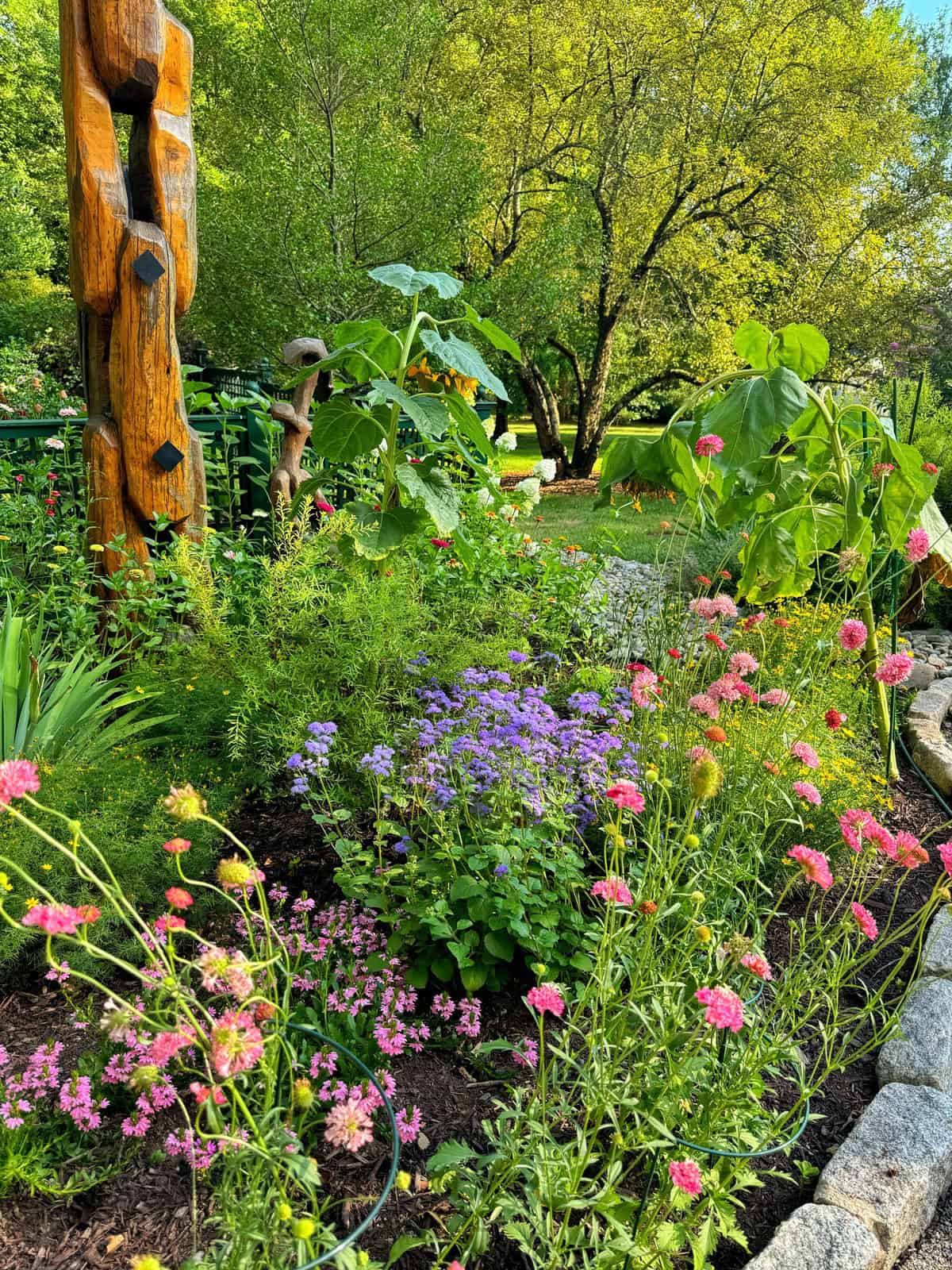 A vibrant garden with various colorful flowers in bloom, including pink and purple blossoms. Large green leaves and a wooden totem pole sculpture with intricate carvings are prominent. Trees and lush greenery fill the background, under a bright, clear sky.