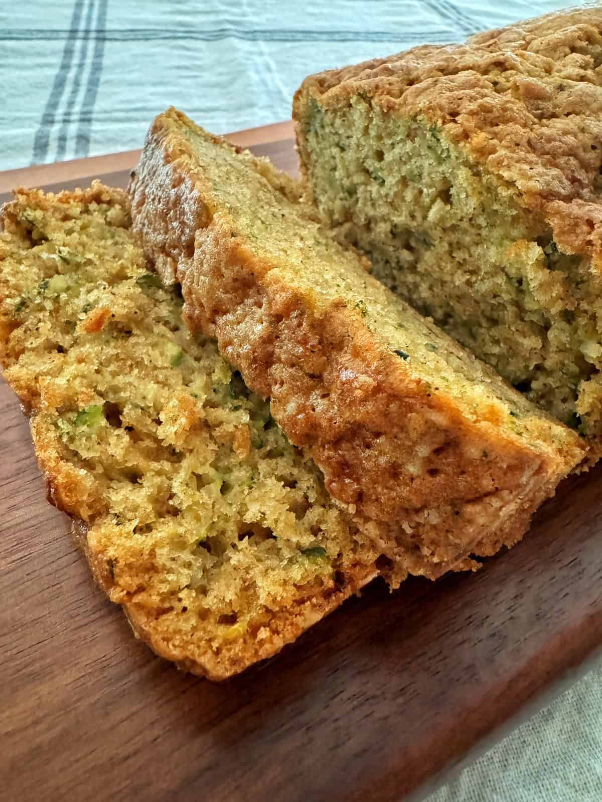 Close-up of sliced zucchini bread on a wooden cutting board. The bread has a golden-brown crust and a moist, light green and yellow interior, with visible pieces of zucchini and other ingredients. A striped cloth in white and blue is in the background.