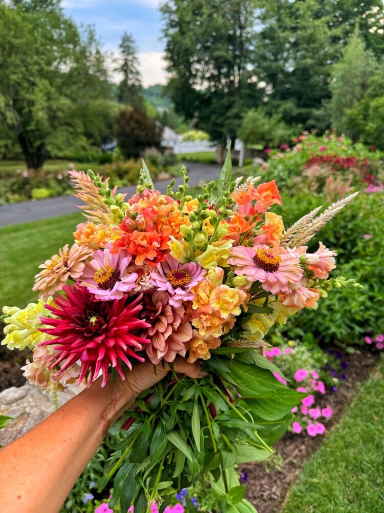 A hand holding a vibrant bouquet of assorted fresh cut flowers, including pink, orange, and red blooms, with a backdrop of a lush garden featuring green grass, trees, and colorful flowers. The scene reflects a serene, sunny day in a well-maintained garden.