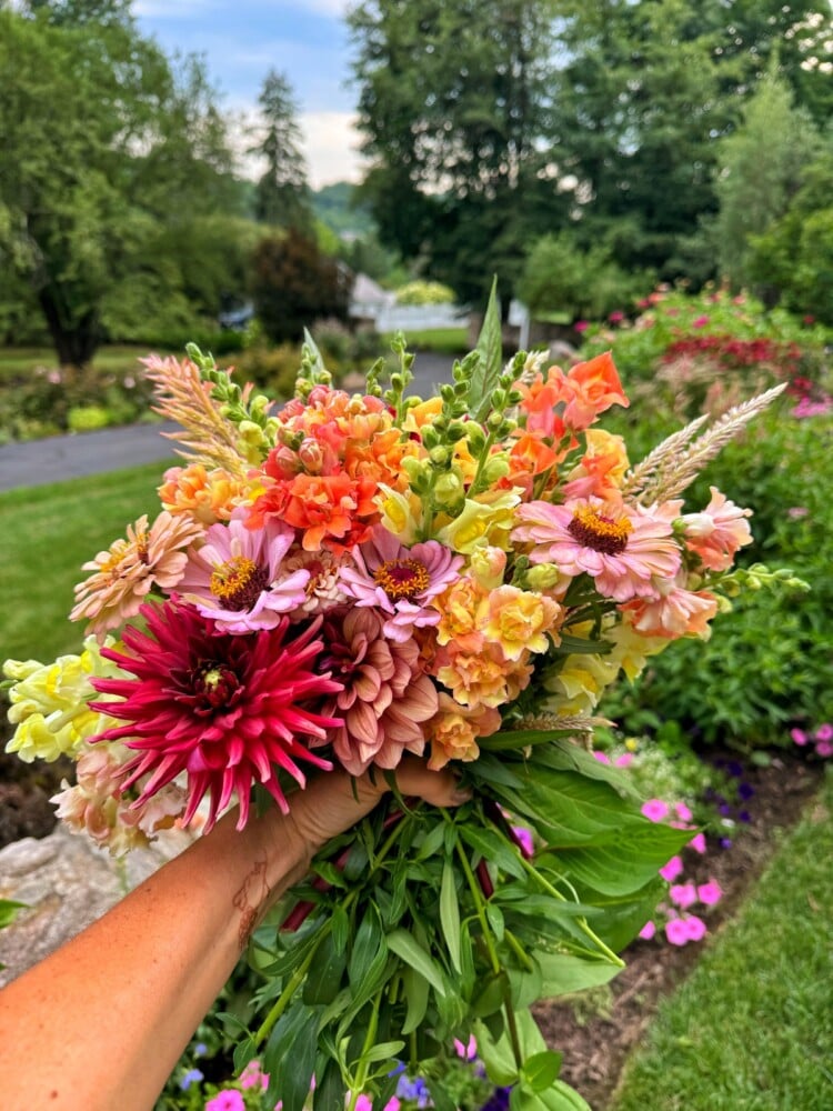A person holds a colorful bouquet of flowers outdoors. The bouquet includes various vibrant blooms in shades of pink, orange, yellow, and red. In the background, a lush garden with green trees, a lawn, and blooming flowers is visible on a bright day.