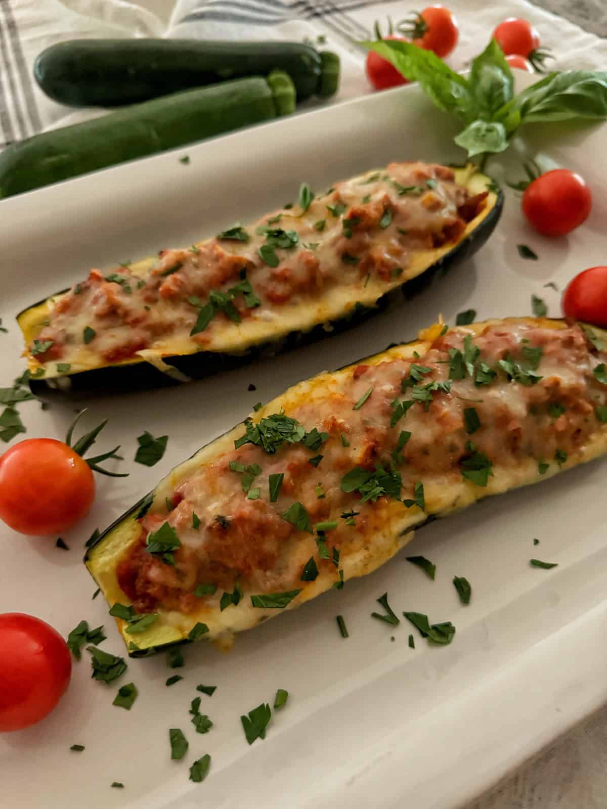Two zucchini boats filled with a mixture of ground meat, tomato sauce, and melted cheese, garnished with chopped parsley, are presented on a white plate. Surrounding the dish are fresh cherry tomatoes, whole zucchinis, and a sprig of basil.