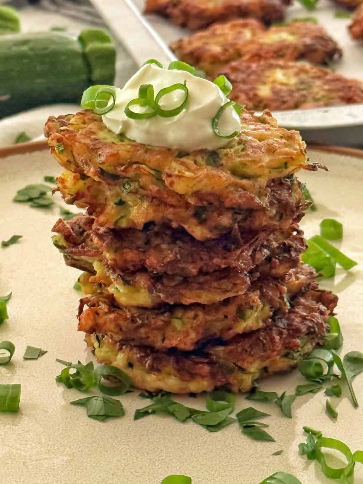 A stack of five golden-brown zucchini fritters topped with a dollop of sour cream and sprinkled with chopped green onions. More fritters are blurred in the background along with whole zucchinis placed nearby, all on a brown-rimmed plate.
