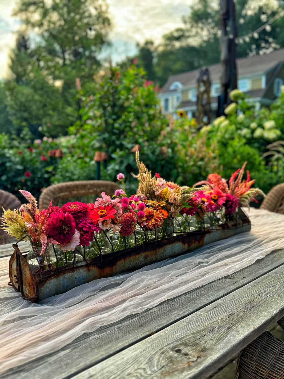 A long wooden table set outdoors with a rustic wooden box centerpiece filled with vibrant mixed flowers, including pink, orange, and red blooms. A white sheer cloth is draped across the table, and greenery with houses is visible in the background.