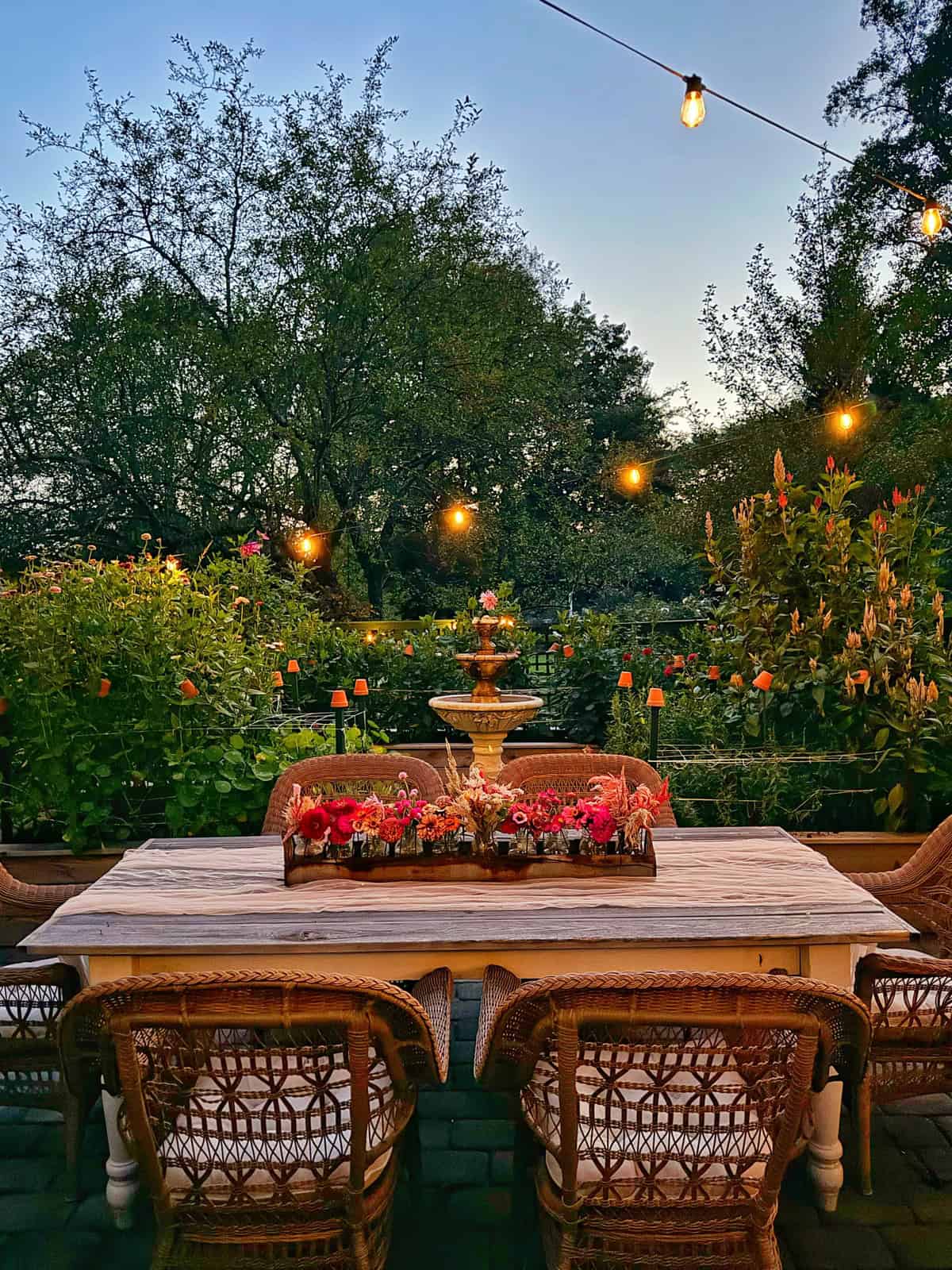 A cozy outdoor dining area at dusk, featuring a wooden table surrounded by wicker chairs. The table is decorated with a floral arrangement, and string lights hang overhead. Lush greenery and blooming flowers form a peaceful garden backdrop.