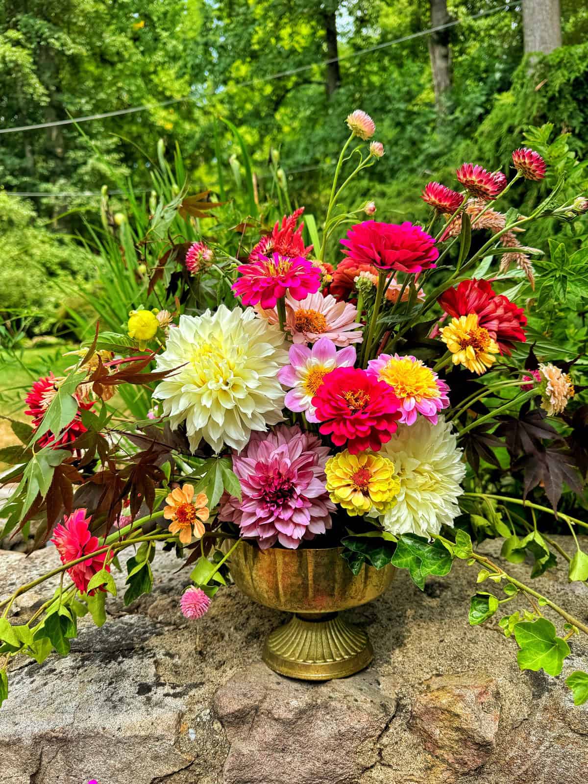 A vibrant floral arrangement in a golden vase sits on a stone surface. The bouquet features an array of brightly colored flowers including pink, red, yellow, white, and orange blooms. In the background, lush green foliage and trees are visible.