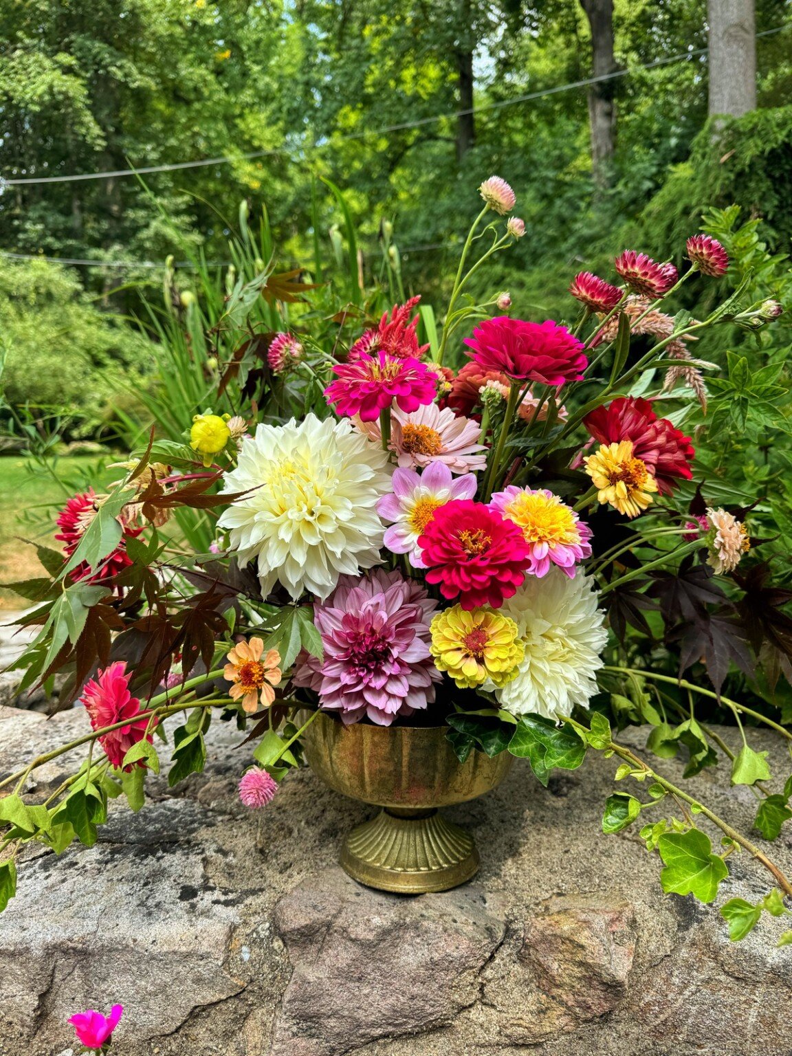 A vibrant bouquet of various flowers, including red, white, pink, and yellow blooms, arranged in a decorative golden vase. The arrangement is set outdoors on a stone surface with a lush, green garden in the background.