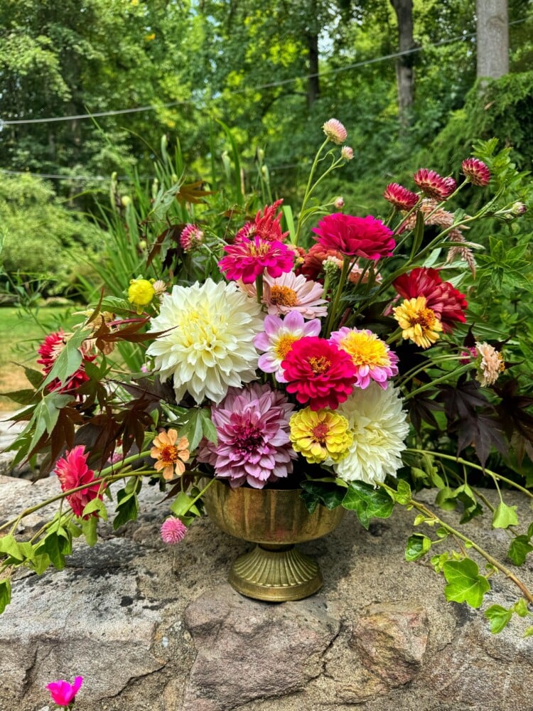 A vibrant bouquet of various flowers, including red, white, pink, and yellow blooms, arranged in a decorative golden vase. The arrangement is set outdoors on a stone surface with a lush, green garden in the background.
