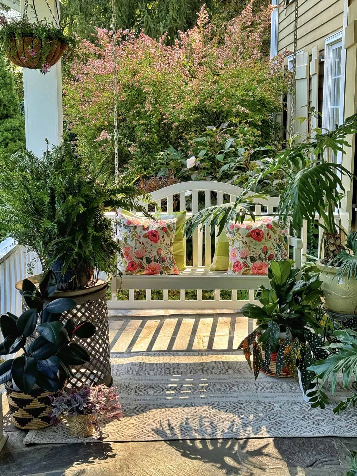 A cozy porch with a white wooden bench adorned with floral cushions. Surrounding the bench are various potted plants, including ferns and green leafy varieties. Pink flowering bushes and a yellow house with siding are visible in the background.