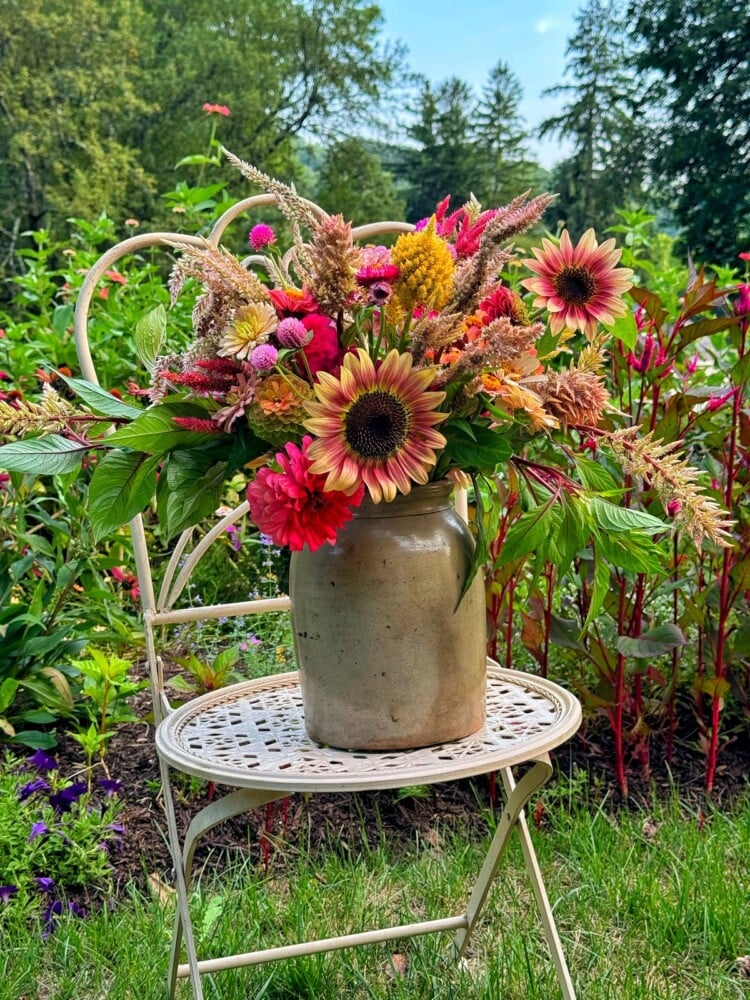 A rustic beige ceramic pot filled with a vibrant arrangement of sunflowers, zinnias, and other colorful flowers sits on a vintage white metal chair in a lush garden, with green foliage and trees in the background.