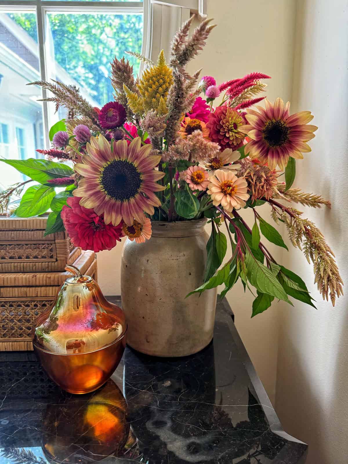 A rustic vase on a dark marble counter holds a colorful bouquet of flowers, including sunflowers, dahlias, and other wildflowers. Next to the vase is a pear-shaped glass container filled with amber liquid, set against a bright window with a partial view outside.