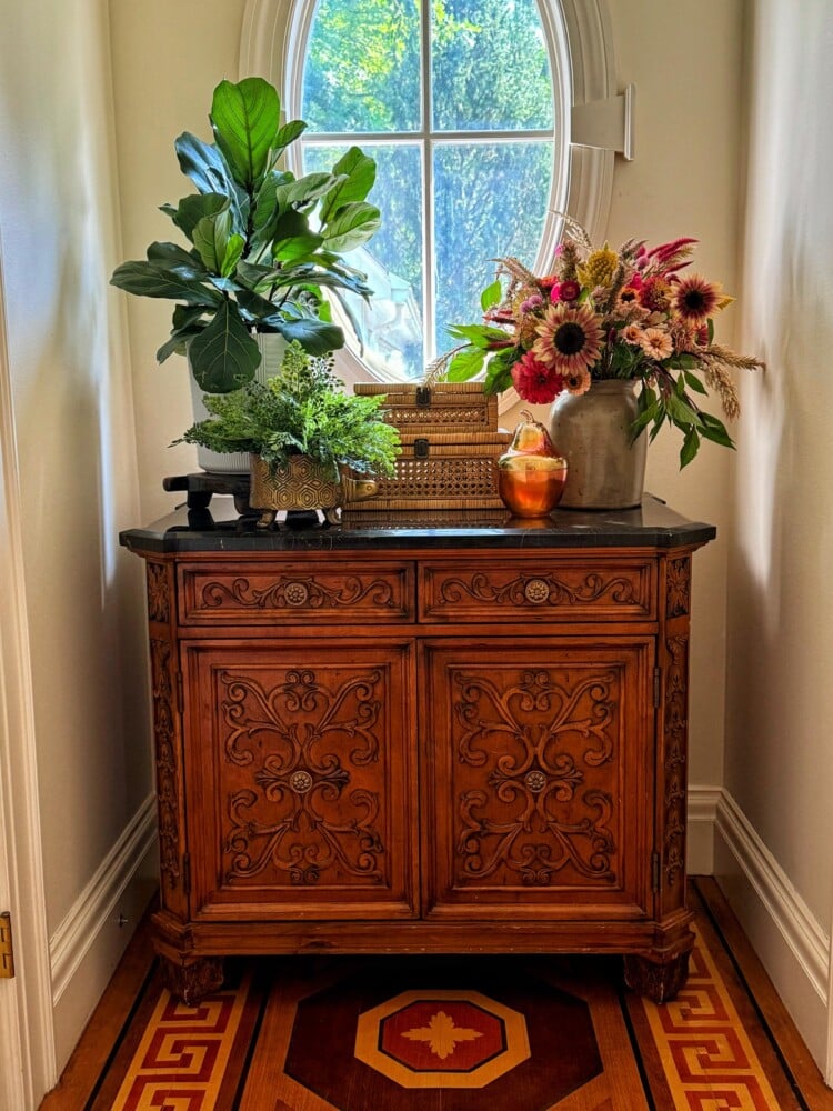 A decorative wooden cabinet sits against a wall under a round window. On top, there are potted plants, a wicker basket, an amber vase, and a bouquet of colorful flowers. The floor features a patterned design in warm colors. Sunlight filters in through the window.
