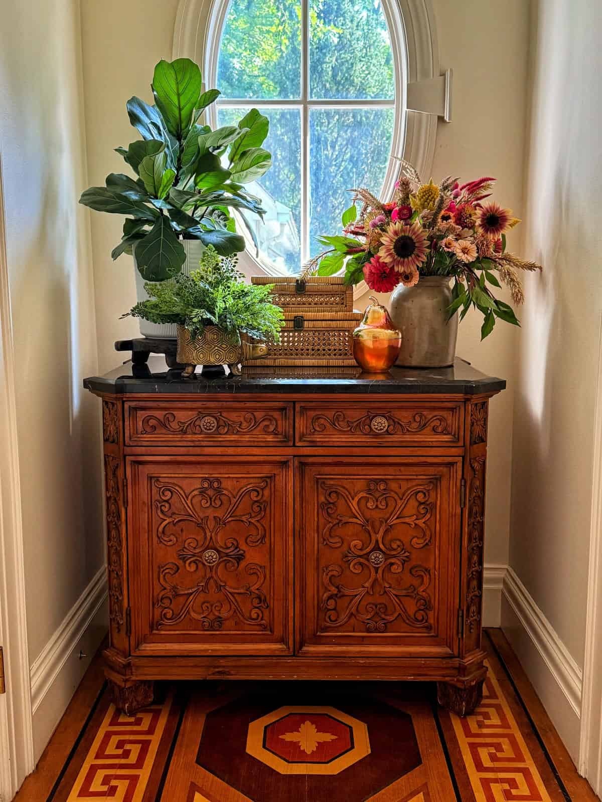 An ornate wooden cabinet with intricate carvings stands in a sunlit hallway. On top are two leafy potted plants, a floral bouquet with sunflowers in a wicker basket, and a small orange vase. A round window above lets in natural light, highlighting the scene.