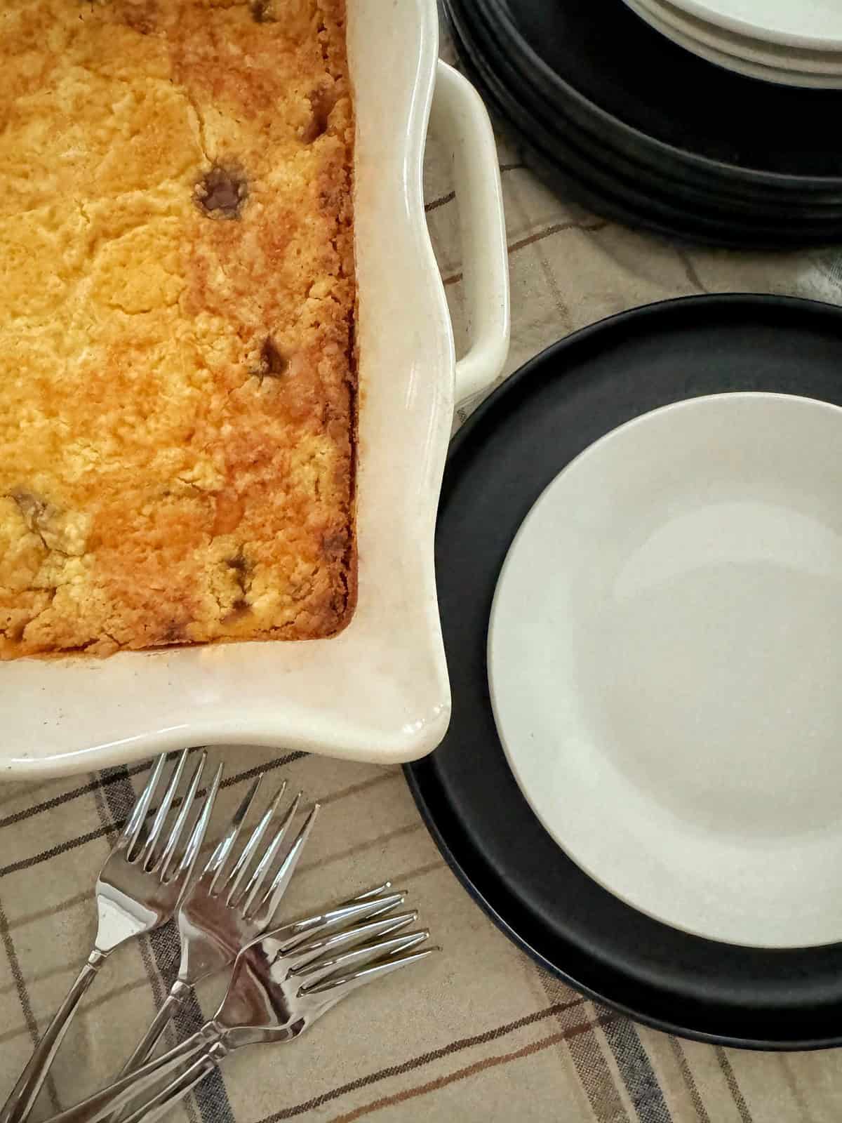A baked casserole dish with a golden crust of easy apple dump cake recipe sits on a plaid tablecloth next to a stack of black-edged plates with white centers. Four forks are placed beside the dish, ready for serving.