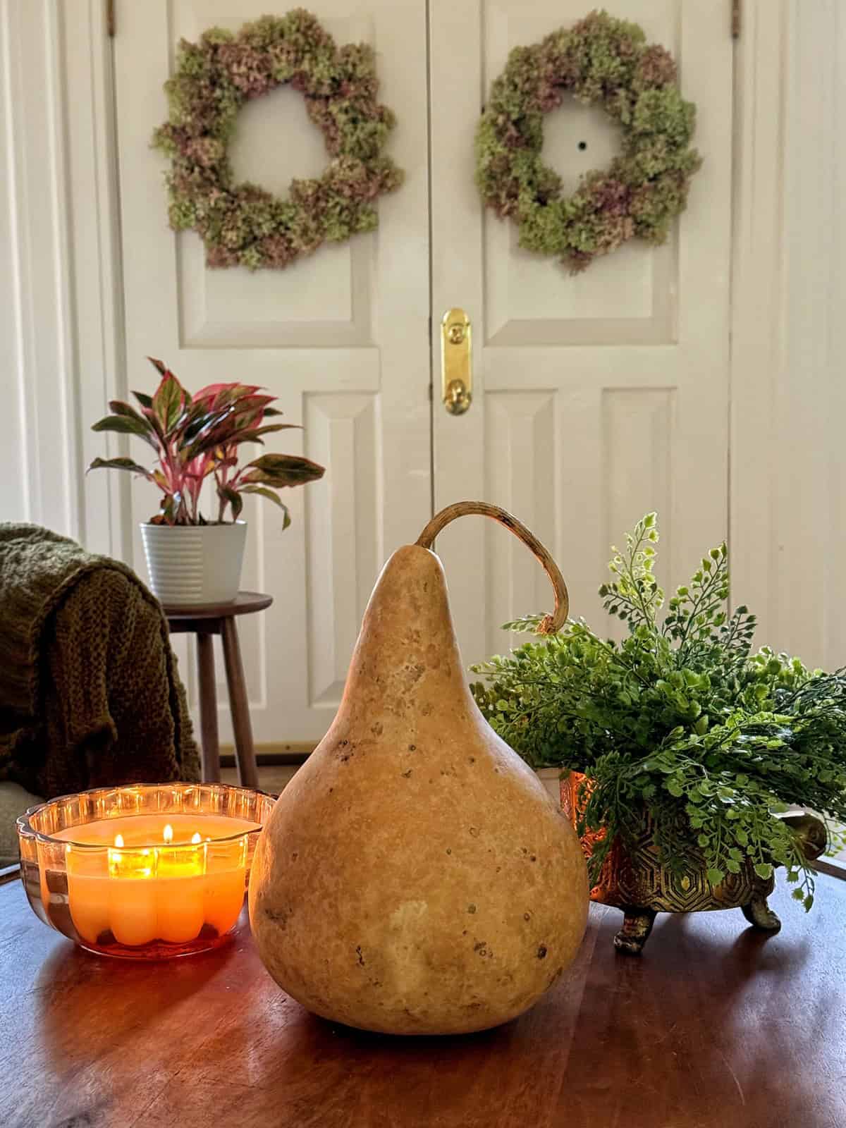 A cozy indoor setting with two green wreaths hanging on a white door. On a wooden table, there's a large dried gourd, a potted plant, a metal planter with greenery, and an amber candle burning with multiple wicks. A chair with a green throw is partially visible.