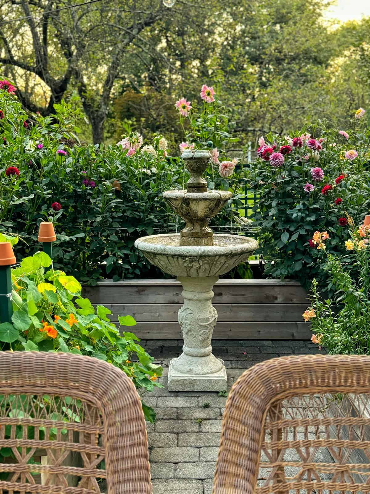 Ornate stone fountain with water cascading from top to bottom tier, surrounded by lush flowering plants and greenery. In the foreground, woven wicker chairs face the fountain, creating a peaceful garden scene under soft daylight with trees in the background.
