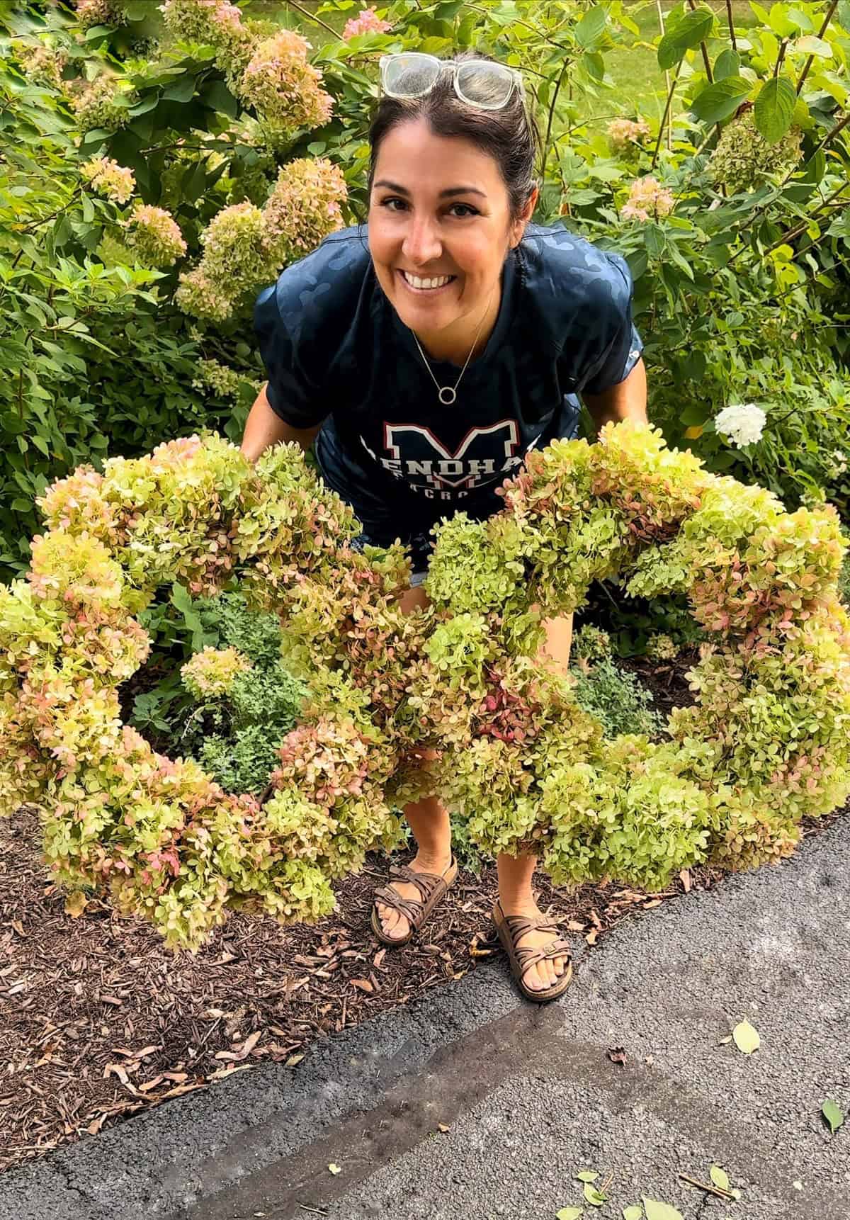 A smiling person wearing a dark blue T-shirt and sandals is standing outdoors, holding a decorative floral arrangement made of greenery and flowers. The background is filled with lush green plants and foliage.
