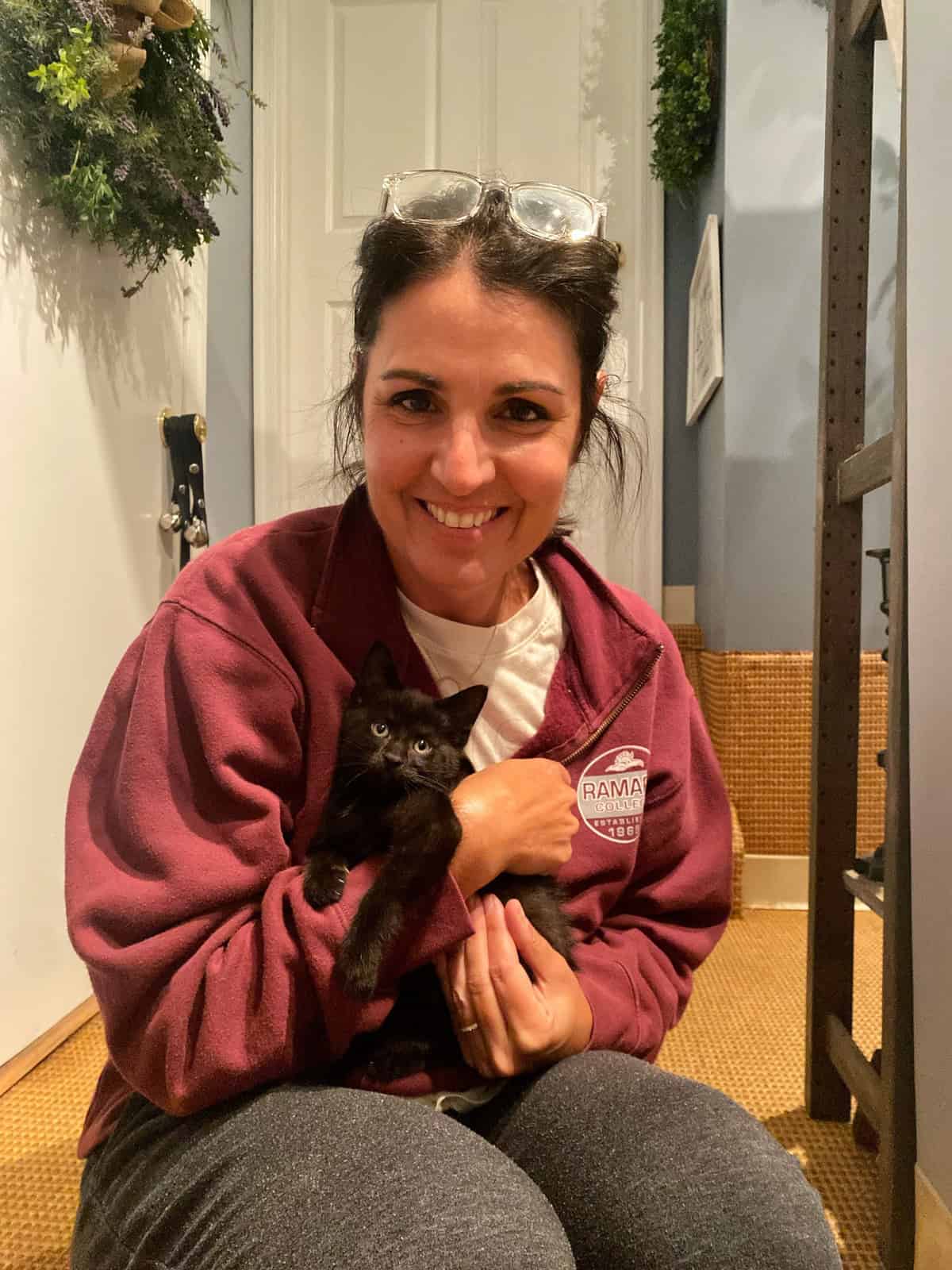 A person (stacy ling) wearing a maroon jacket and gray pants is sitting on a carpeted floor, smiling and holding a small black kitten. They have glasses resting on their head and are sitting near a white door with a wreath hanging on it.
